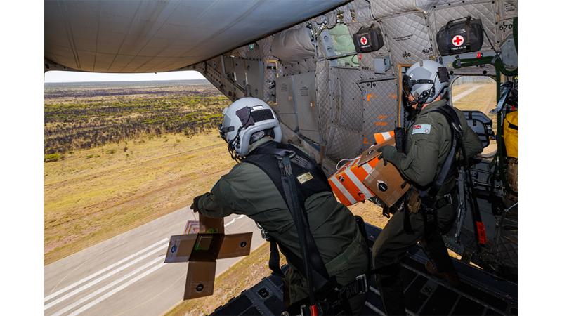 Two military members drop cardboard boxes out of an aircraft in flight.