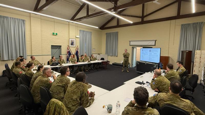 People in uniform sit at tables arranged in a U-shape while a presenter speaks at the front of the room.