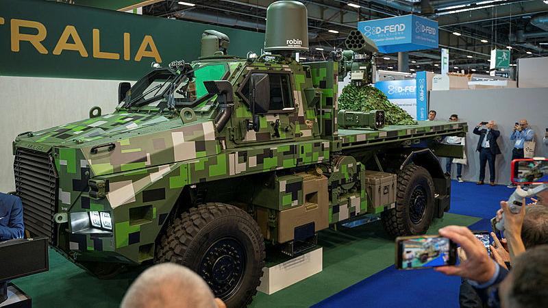 A green camouflage Army vehicle sits on display in an indoor area. People observe and take photos.