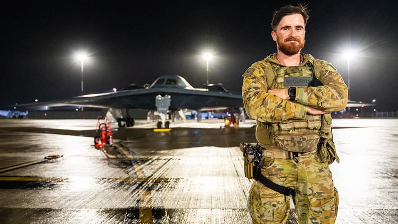 A military member stands in front of a stationary aircraft at night.