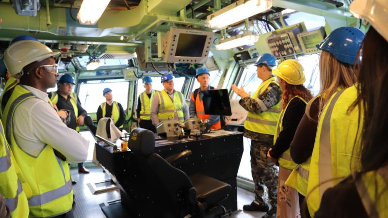 A group of people in high-visibility vests and hard hats stand in a ship’s bridge.