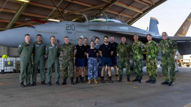 A group of aviators from Australia, Sweden and Finland stand with their backs to a jet aircraft and smile for the camera.