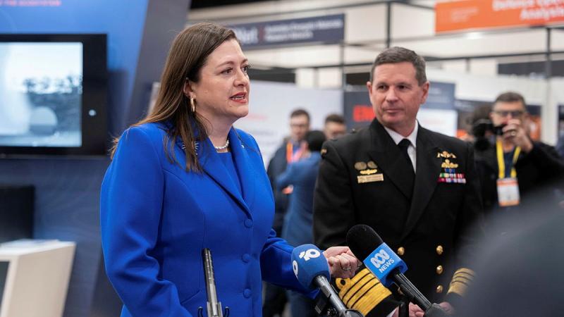 A woman in a blue suit and a man in a Navy uniform speak to people with microphones.