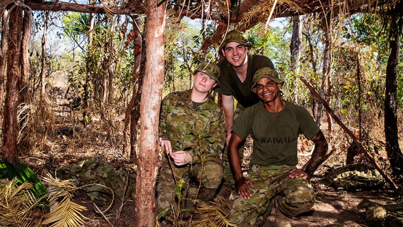 Three military personnel in a man-made shelter in the outdoors.