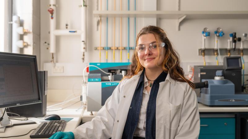 A woman in a lab coat and protective eyewear sits at a laboratory workstation.