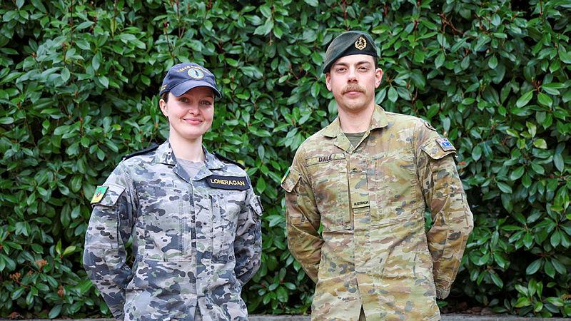 A Navy member and an Army member stand side by side in front of foliage.