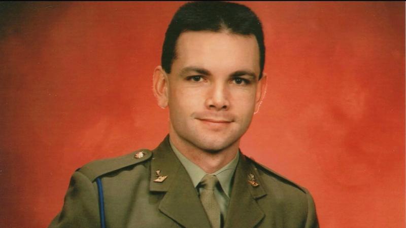 A man in formal Army uniform sits in front of a red backdrop.
