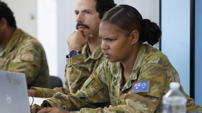 A woman in uniform sits at a table using a laptop, with a man sitting next to her.