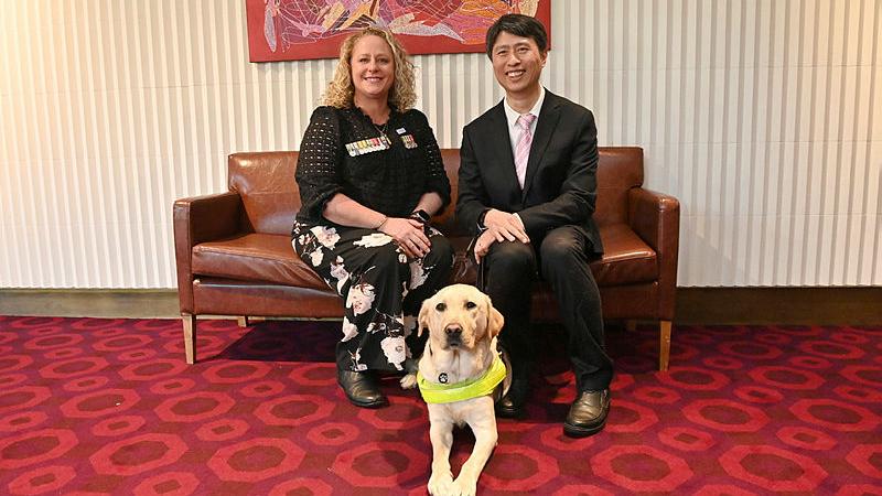 A woman sits on a brown lounge with a man. A Labrador dog sits in front and between them on the floor.