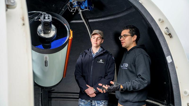 A person in an Air Force uniform and someone in plain clothes stand with a telescope.