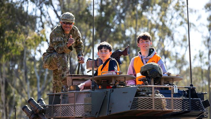 A man in Army uniform and two students in orange high-vis outfits stand on top of a tank.