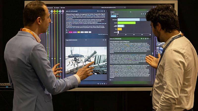 Two men stand looking at a screen which displays text and images.