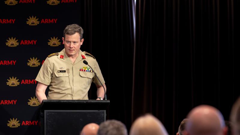 A man in Army uniform stands behind a podium and speaks to an audience.