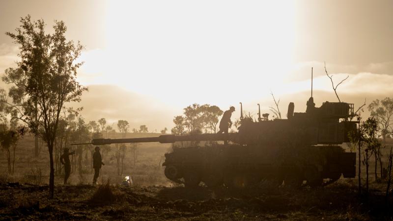Army soldiers stand near and atop a vehicle with a blazing sunset as its background.