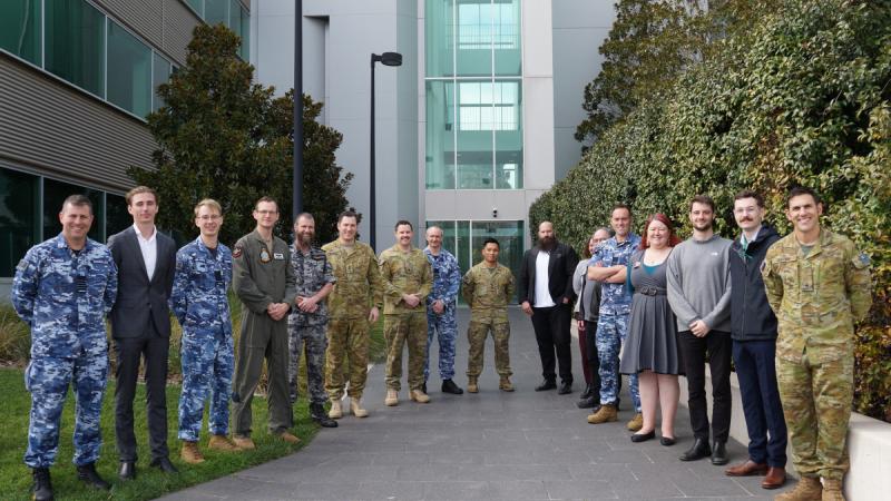 A group of military personnel stand outside a building in a circle and smile at the camera.
