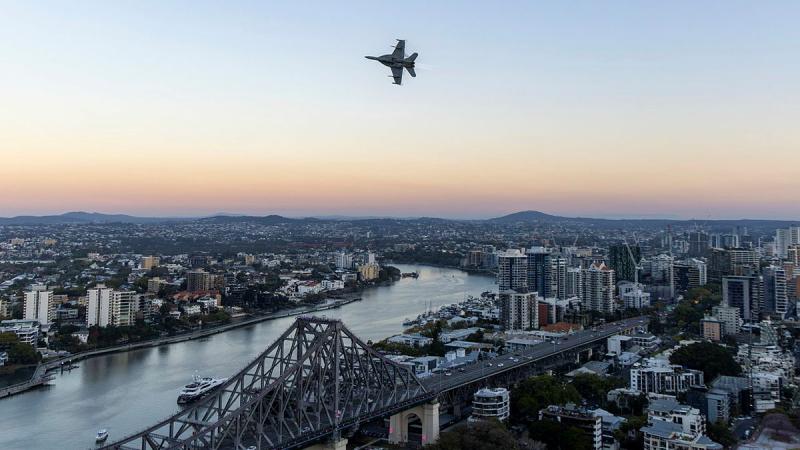 A jet flies over a cityscape and river at dusk.