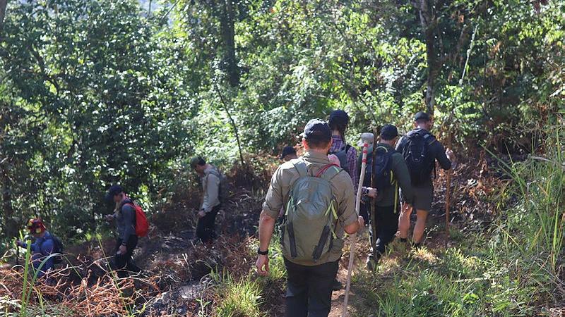 A group of people with backpacks walk along a dirt track with greenery surrounding it.