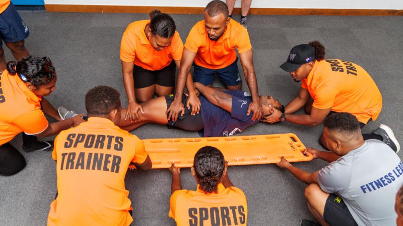 A group of people kneel around a spinal board while one person lies on the ground beside it.