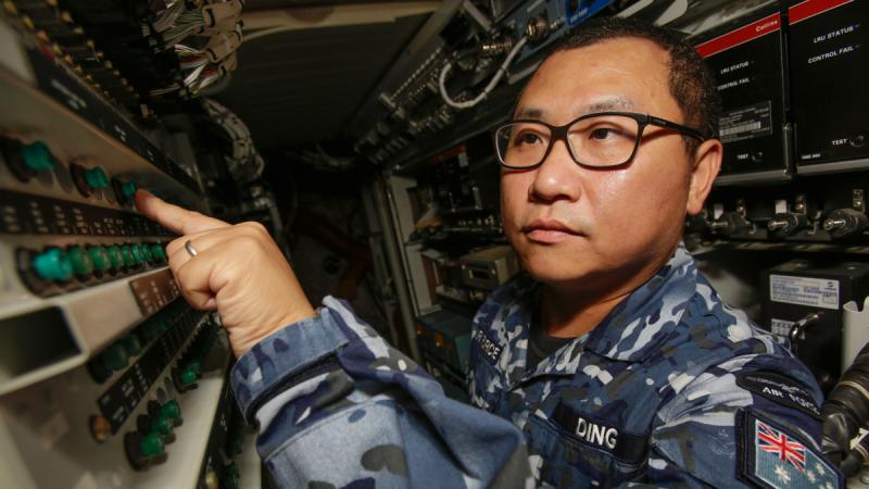 A man in Air Force uniform presses a green button among rows of controls in a confined space.