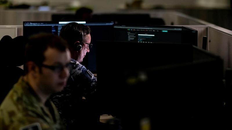 Two people in military uniforms work with computer screens in a darkened room.