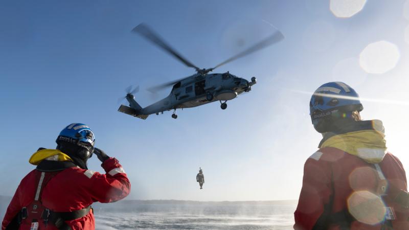 A person dangles from a winch below a helicopter above the water.