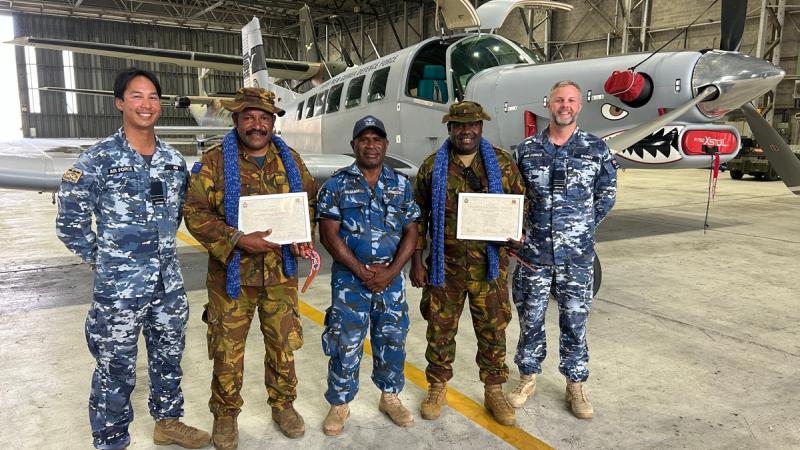 Five military men stand in front of an aircraft, two holding certificates.