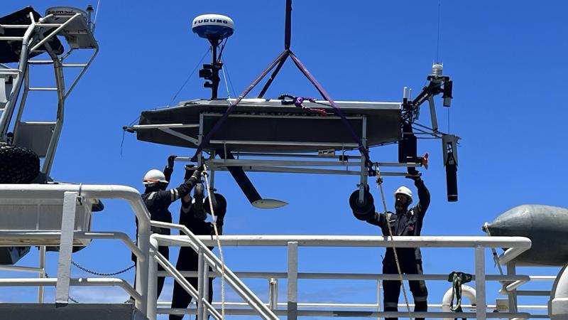 A vessel is hoisted and hangs by a cable as three people steady it at opposite ends.