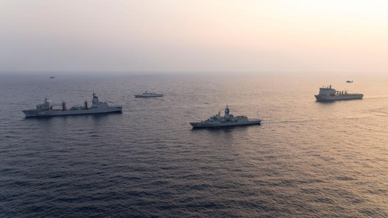 Four ships sail in formation at sea with two helicopters flying above them.