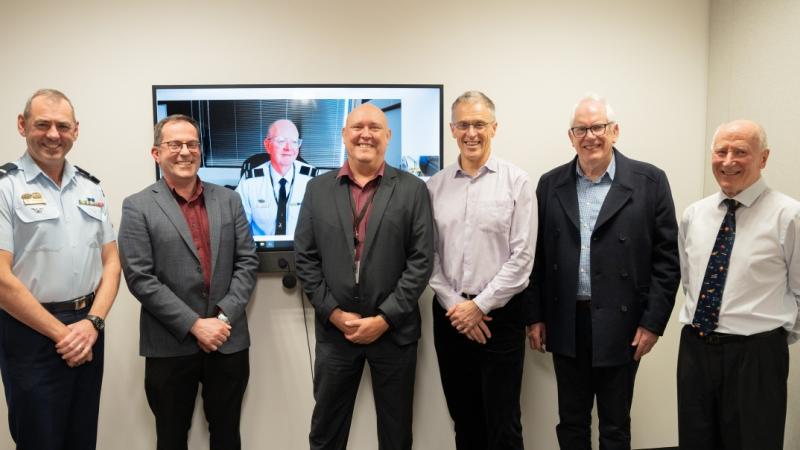 Group photo of AIRCDRE James Badgery (current DG DASA); AIRCDRE Joseph Medved; AIRCDRE Jason Agius (Screen); AIRCDRE James Hood; AIRCDRE Terrence Saunder; AIRCDRE David Tindal (Retd); and Air Vice-Marshal Noel Schmidt (Retd), standing in an office in front of a television screen. 