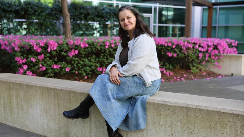 A woman sits cross-legged on a low concrete wall with pink flowers blooming in the garden behind her.