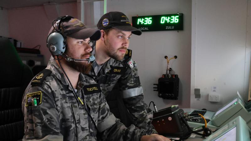 A man in Navy uniform sits at a workstation while another looks at the screen over his shoulder.