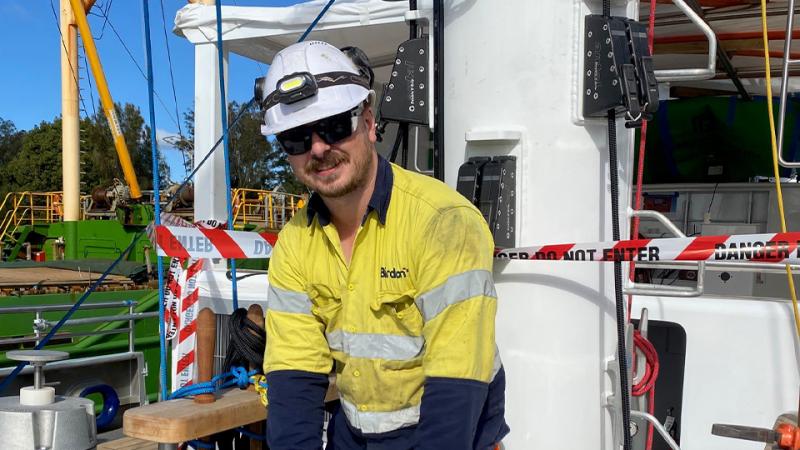 A man in a high-visibility shirt and hard hat stands on the deck of a ship.